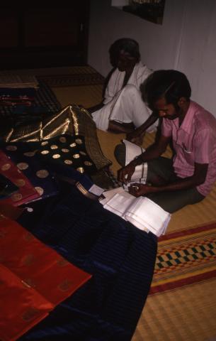 An old woman and a man are looking at spreaded silk clothes (saree, vetti) on a mat